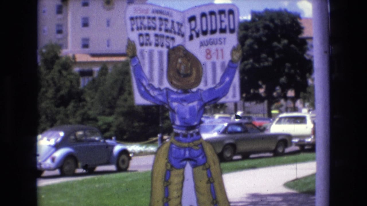 1973 Colorado Springs frame with a colorful Pikes Peak rodeo advertisement, illustrated cowboy figure, parked cars, trees, lawn, and a building in the background.