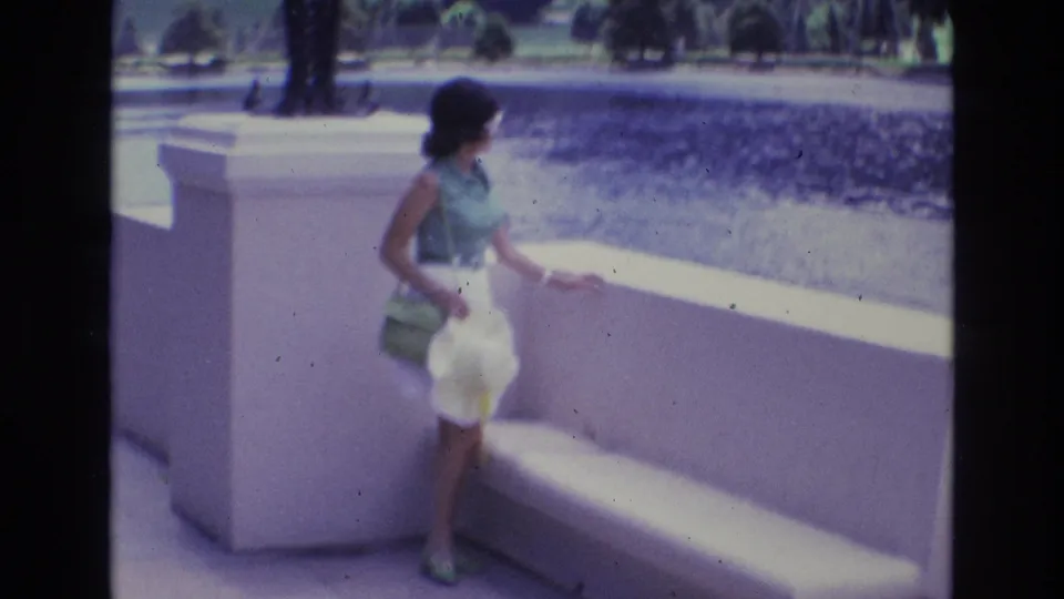 1973 Colorado Springs home-movie frame showing a woman in sunglasses, sleeveless green top, and light skirt standing beside a concrete railing overlooking water.