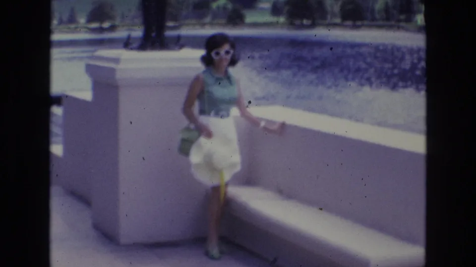 1973 Colorado Springs frame of a woman in sunglasses and 1970s clothing standing at a concrete railing, with water, lawn, trees, and walkway visible behind her.