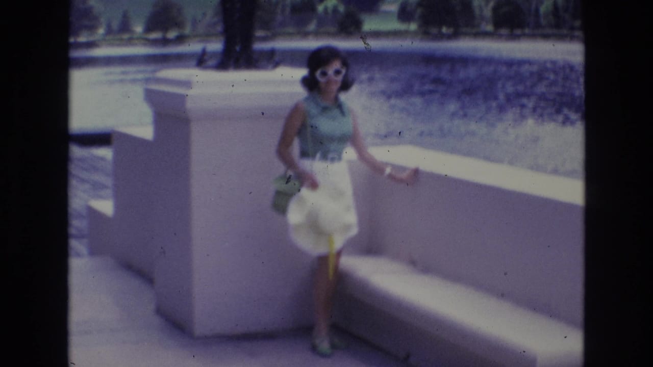 Adult woman in white sunglasses, green sleeveless top, and white skirt poses beside a white concrete bench in 1973 Colorado Springs home-movie footage.