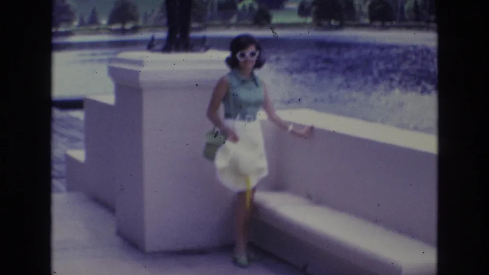 Adult woman in white sunglasses, green sleeveless top, and white skirt poses beside a white concrete bench in 1973 Colorado Springs home-movie footage.