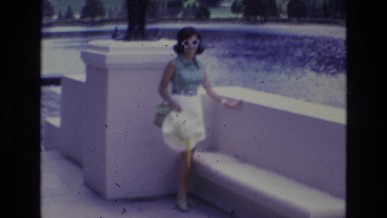 Adult woman in a 1970s outfit stands near a low white wall and bench, holding a wide-brim hat while facing the home-movie camera.