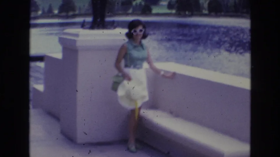 Adult woman in a 1970s outfit stands near a low white wall and bench, holding a wide-brim hat while facing the home-movie camera.