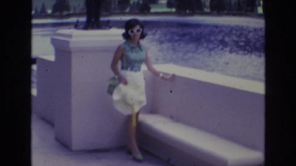 Adult woman wearing sunglasses and a white skirt poses by a white concrete bench, with trees and open grounds visible behind her in the soft 8mm frame.