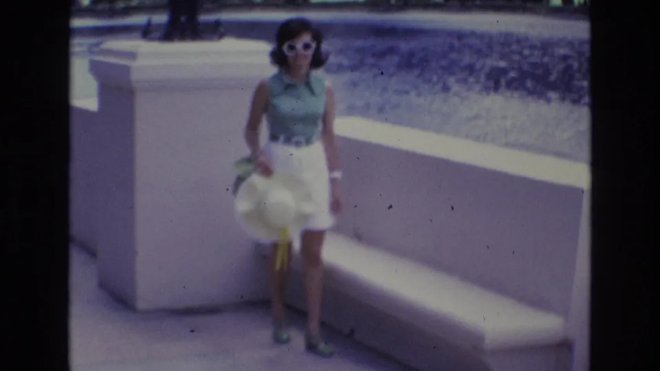 Adult woman in sunglasses stands close to a white bench and low wall, holding a hat with a small green shoulder bag visible at her side.