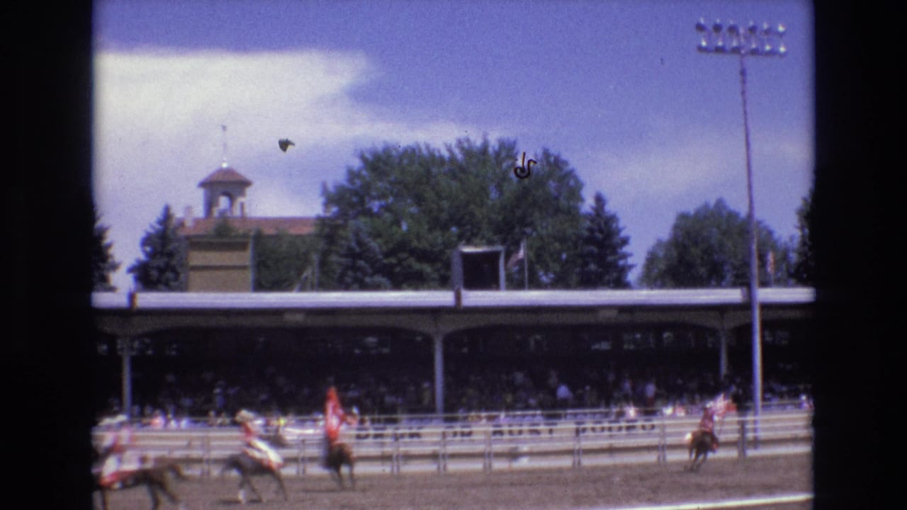 In supplied 1973 Colorado Springs footage, mounted riders pass a dirt rodeo arena while spectators sit beneath a covered grandstand and a cupola-topped structure rises behind trees.