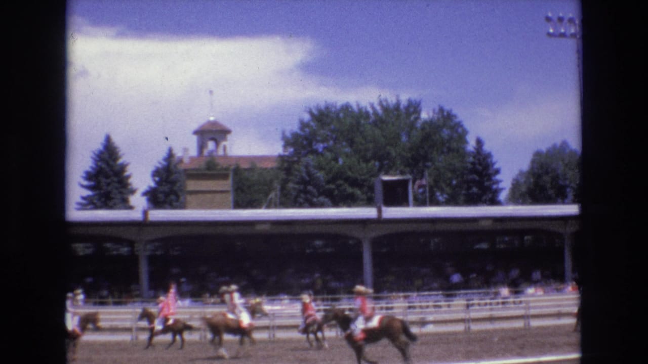 In the 1973 Colorado Springs rodeo scene, multiple riders in colorful attire move across the dirt arena with a shaded spectator stand, trees, and arena railings behind them.