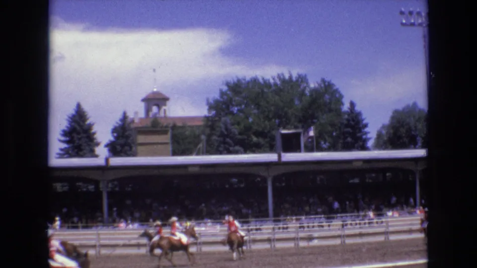The home-movie frame shows rodeo riders moving along the arena rail in Colorado Springs, with spectators under a long roof and a cupola-topped building visible above the trees.