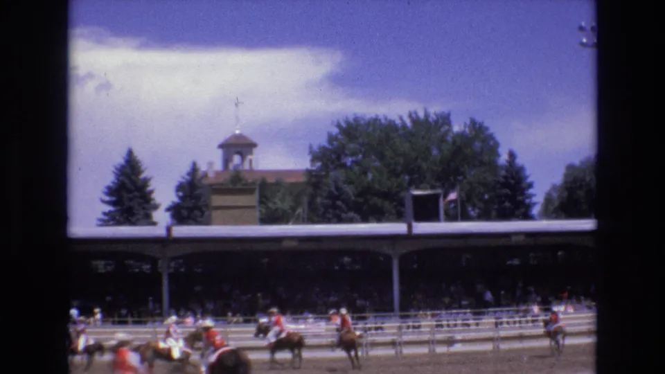 At the end of the clip, riders and horses appear across the Colorado Springs rodeo arena, framed by railings, shaded spectators, trees, and a small cupola structure in the background.