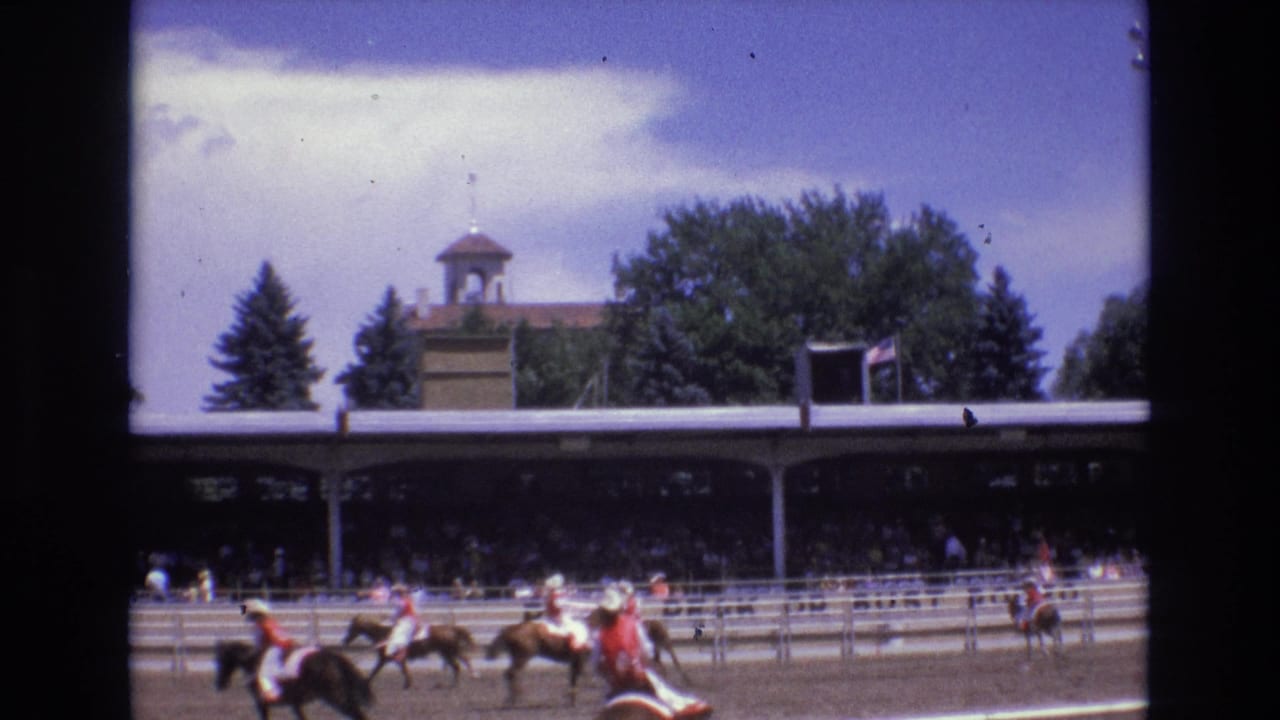 In the supplied 1973 Colorado Springs home-movie footage, jockeys on horseback race along a dirt track in front of a covered grandstand and a cupola-topped building.