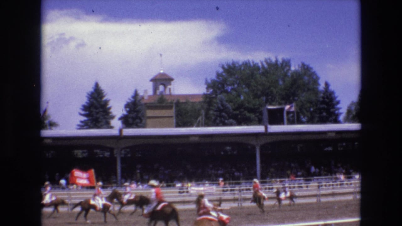 Riders on horses move along the rail while spectators watch from a shaded grandstand.