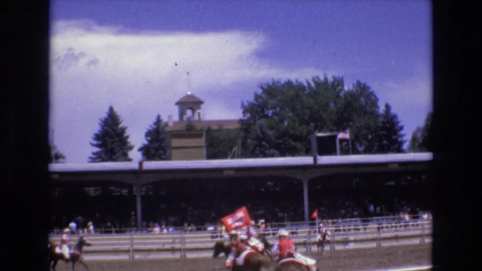 The home-movie frame shows jockeys riding past a red sign or banner beside the grandstand, with the cupola-topped building and trees beyond the track.