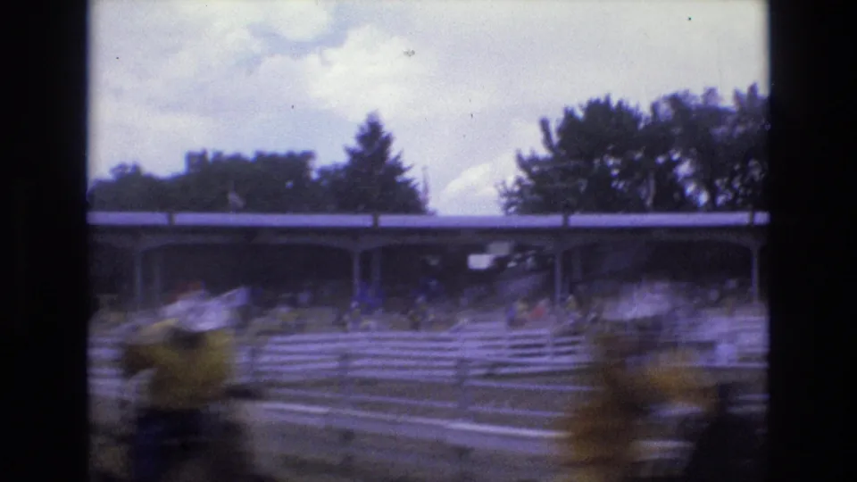 Fast-moving horses blur across the foreground as the camera follows the race past the rail fence, grandstand, trees, and cloudy sky.