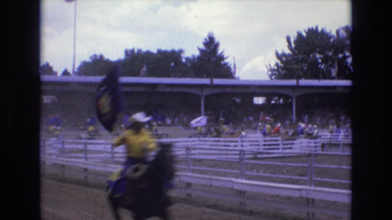 In 1973 Colorado Springs, mounted rodeo riders in yellow shirts and cowboy hats carry flags past white fencing and shaded spectator stands.