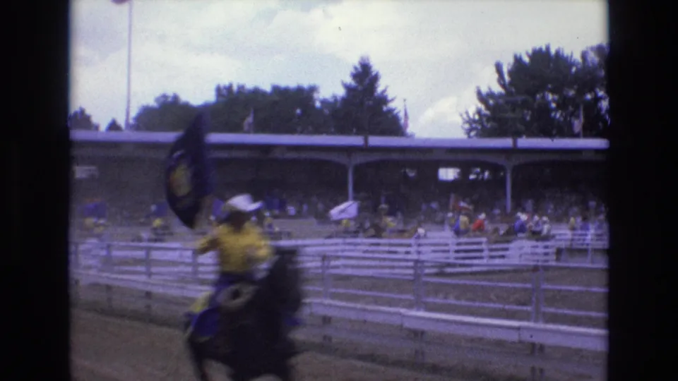 In 1973 Colorado Springs, mounted rodeo riders in yellow shirts and cowboy hats carry flags past white fencing and shaded spectator stands.