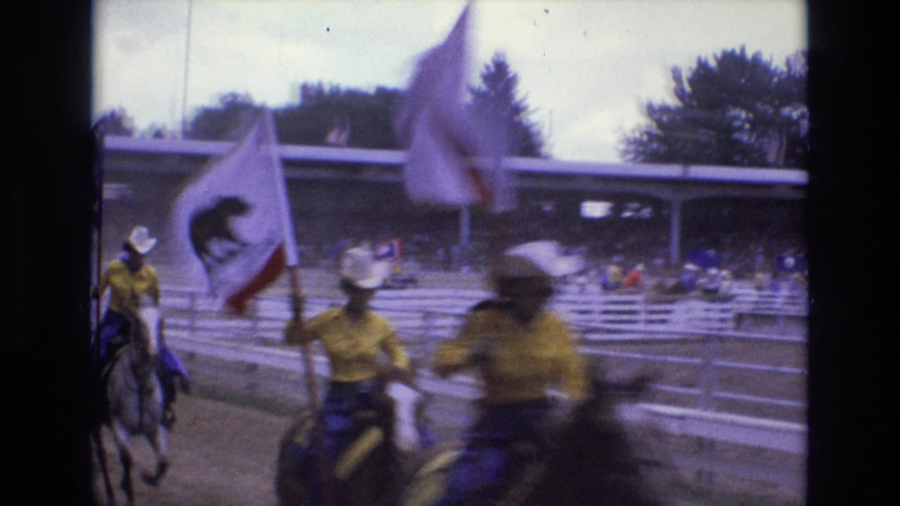 Mounted riders in a Colorado Springs rodeo procession carry large state flags past the camera, with arena rails and spectators behind them.
