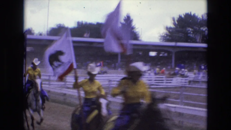 Mounted riders in a Colorado Springs rodeo procession carry large state flags past the camera, with arena rails and spectators behind them.