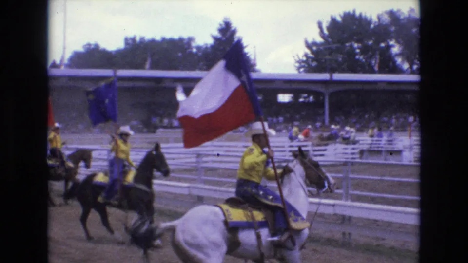 A rider in yellow carries a Texas flag on horseback in the Colorado Springs rodeo arena, with other flag-bearing riders and spectators visible behind.