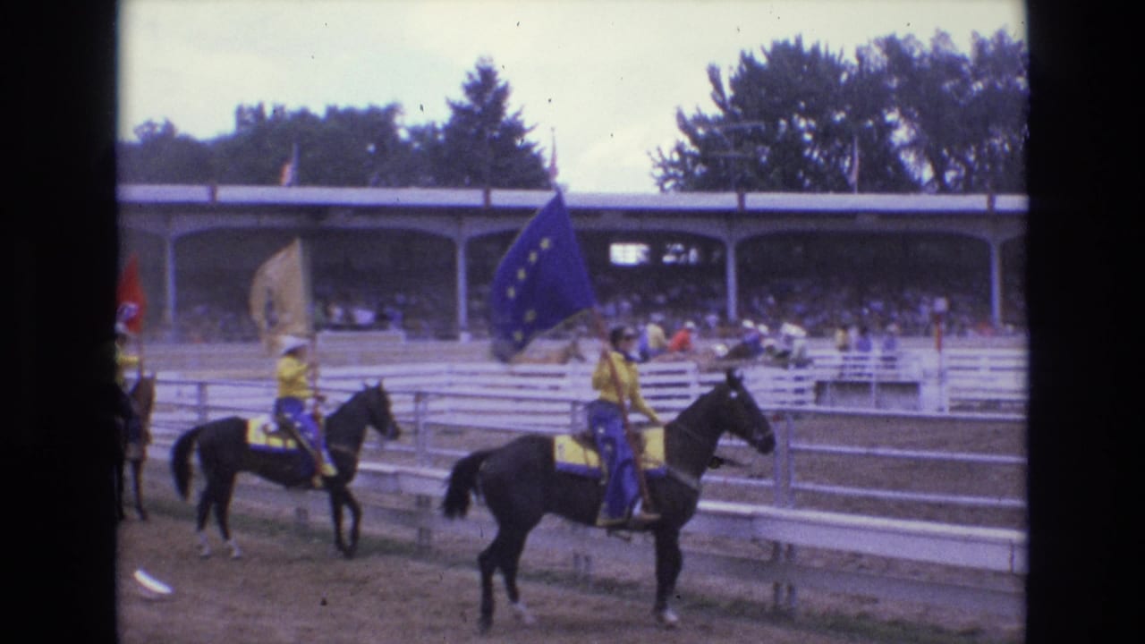 A line of rodeo riders on horseback carries flags across the arena with spectators seated under a covered stand behind them.