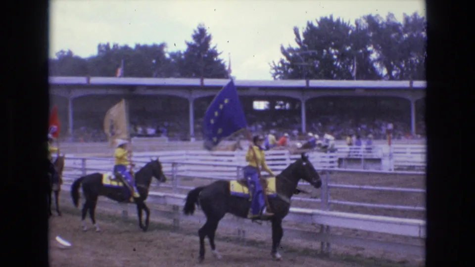 A line of rodeo riders on horseback carries flags across the arena with spectators seated under a covered stand behind them.