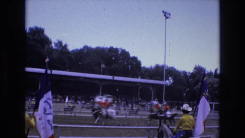 From the spectator-side view, a flag bearer in a white hat stands near the rail while other riders move quickly across the rodeo arena.