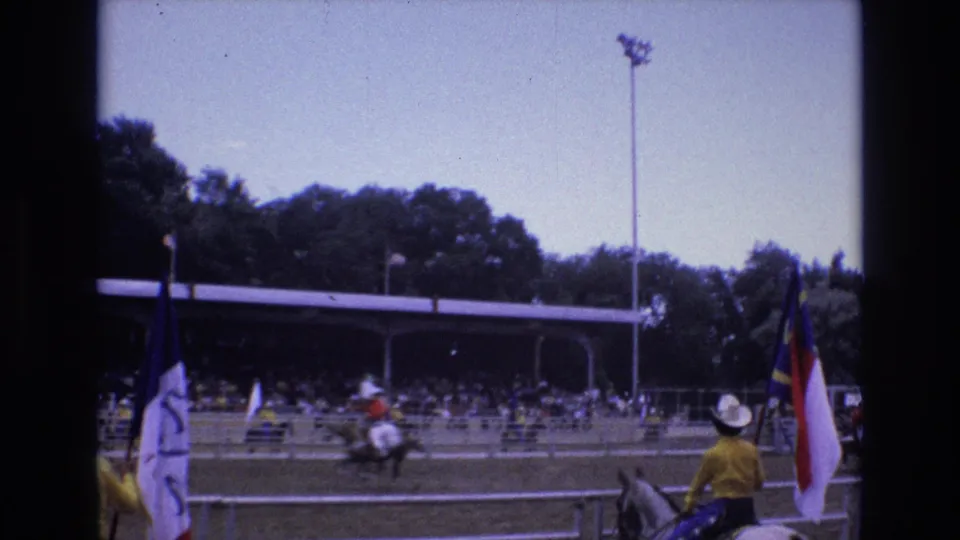 A mounted rider holding a red, white, and blue flag stands near the foreground while other horseback riders cross the arena before the covered stands.