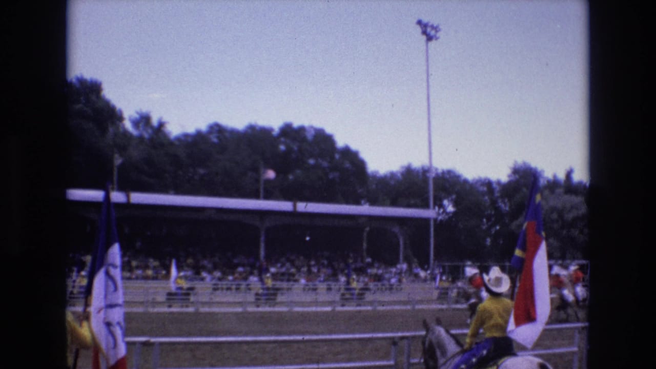 In 1973 Colorado Springs footage, mounted riders carrying flags pass an outdoor arena grandstand while spectators sit under a covered bleacher roof.