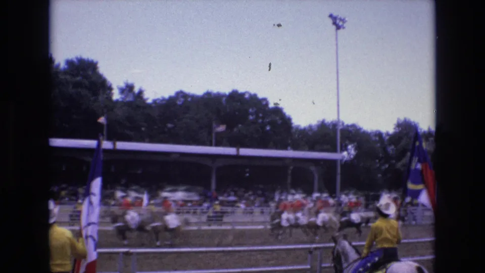 Mounted riders in western attire move across the Colorado Springs rodeo arena while flag bearers and a crowd in covered bleachers remain visible in the background.