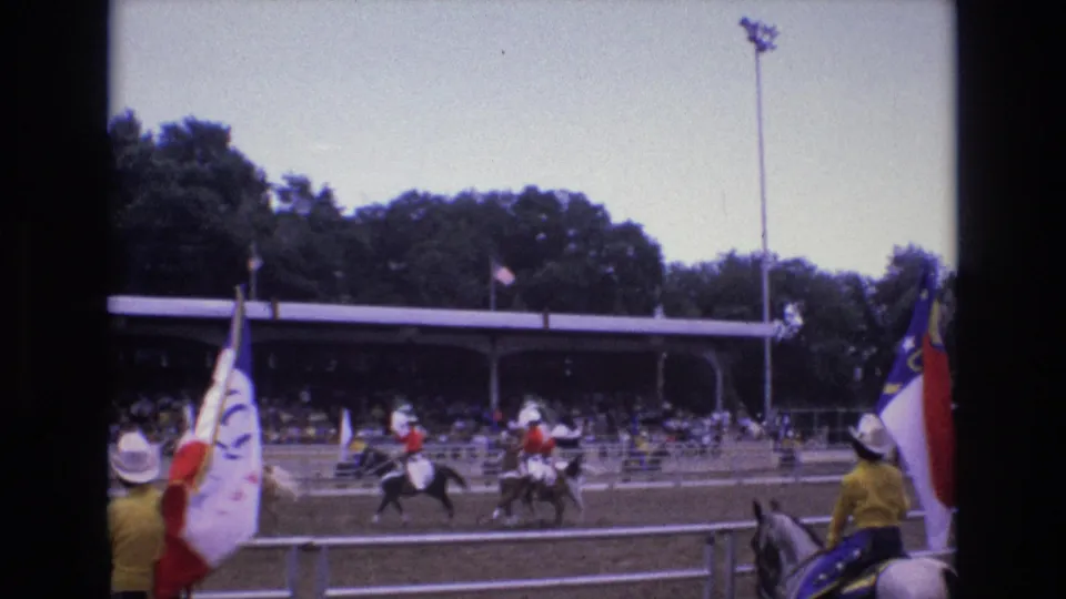 Two mounted riders pass through the rodeo arena in the 1973 Colorado Springs home-movie footage, with flag bearers, spectators, trees, and a tall light pole behind them.