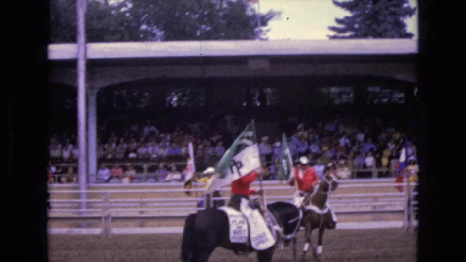 A closer view shows horseback riders with a green, white, and red flag moving along the arena rail, with spectators seated in the covered grandstand behind them.
