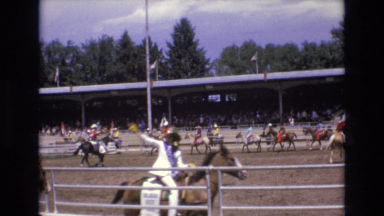 1973 Colorado Springs rodeo footage showing a rider in white western attire on horseback in the foreground, with other mounted riders and spectators under a covered pavilion behind the arena.