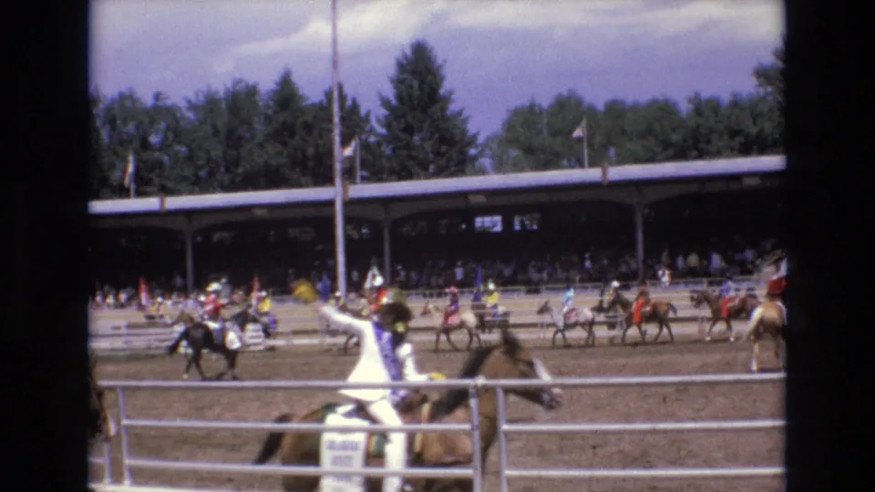 1973 Colorado Springs rodeo footage showing a rider in white western attire on horseback in the foreground, with other mounted riders and spectators under a covered pavilion behind the arena.