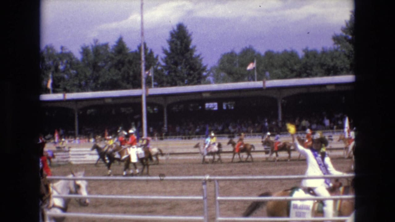 1973 Colorado Springs home-movie frame of mounted rodeo riders spread across a dirt arena, with a foreground fence, covered grandstand, flags, and trees beyond the pavilion.