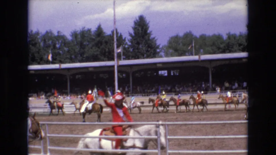 1973 Colorado Springs rodeo frame showing a rider in red western attire on a light-colored horse near the metal fence, with more riders and a covered spectator pavilion in the background.