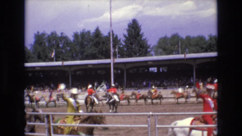 1973 Colorado Springs rodeo footage with multiple riders across the dirt arena, foreground horses and riders at the fence, a shaded spectator pavilion, flags, trees, and a cloudy sky.