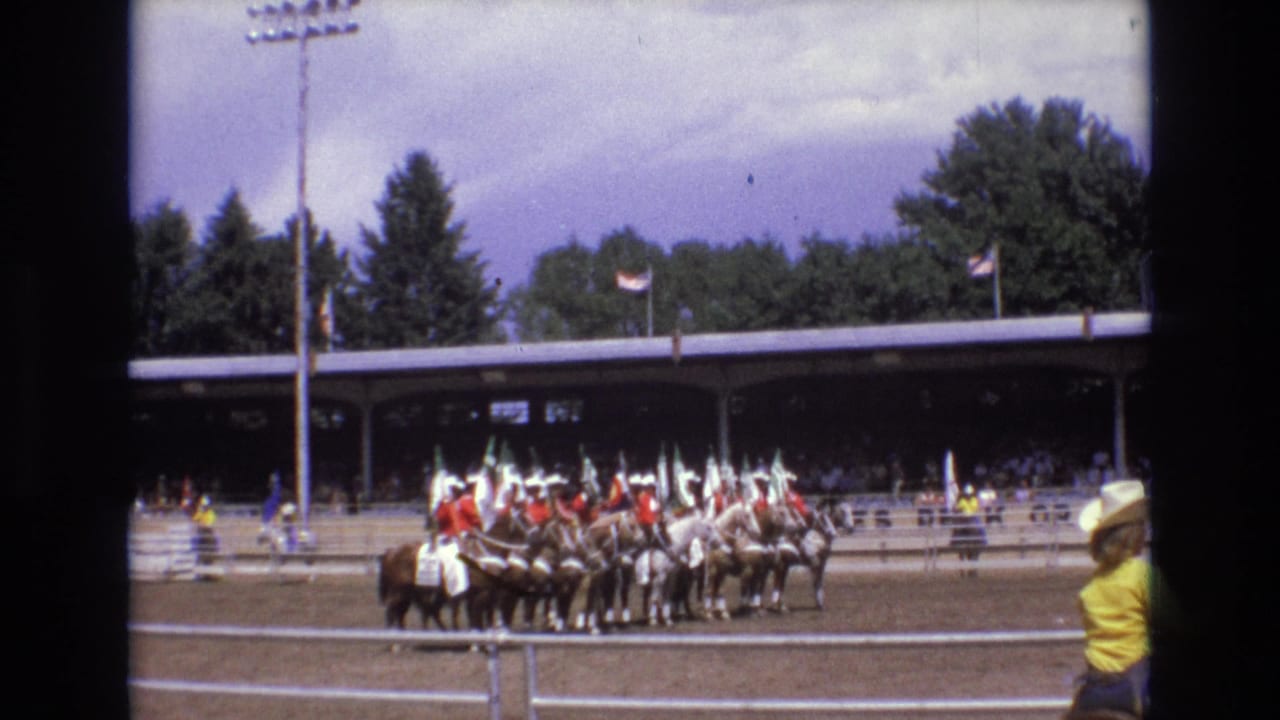 1973 Colorado Springs rodeo footage shows mounted riders with flags grouped in a dirt arena while spectators sit under a covered grandstand.