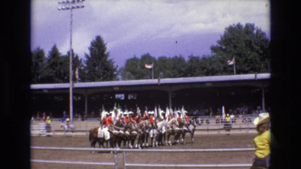 1973 Colorado Springs rodeo footage shows mounted riders with flags grouped in a dirt arena while spectators sit under a covered grandstand.