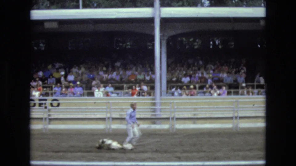 Near the end of the 1973 rodeo clip, the roper stands beside the calf in the arena with crowded bleachers and partial signage behind him.
