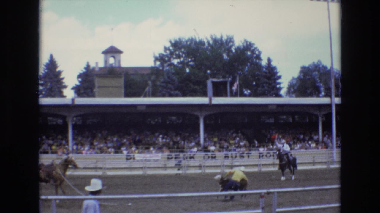 1973 Colorado Springs rodeo footage showing cowboys on horseback, a small bull or calf near the rail, and a crowded covered grandstand behind the arena fence.