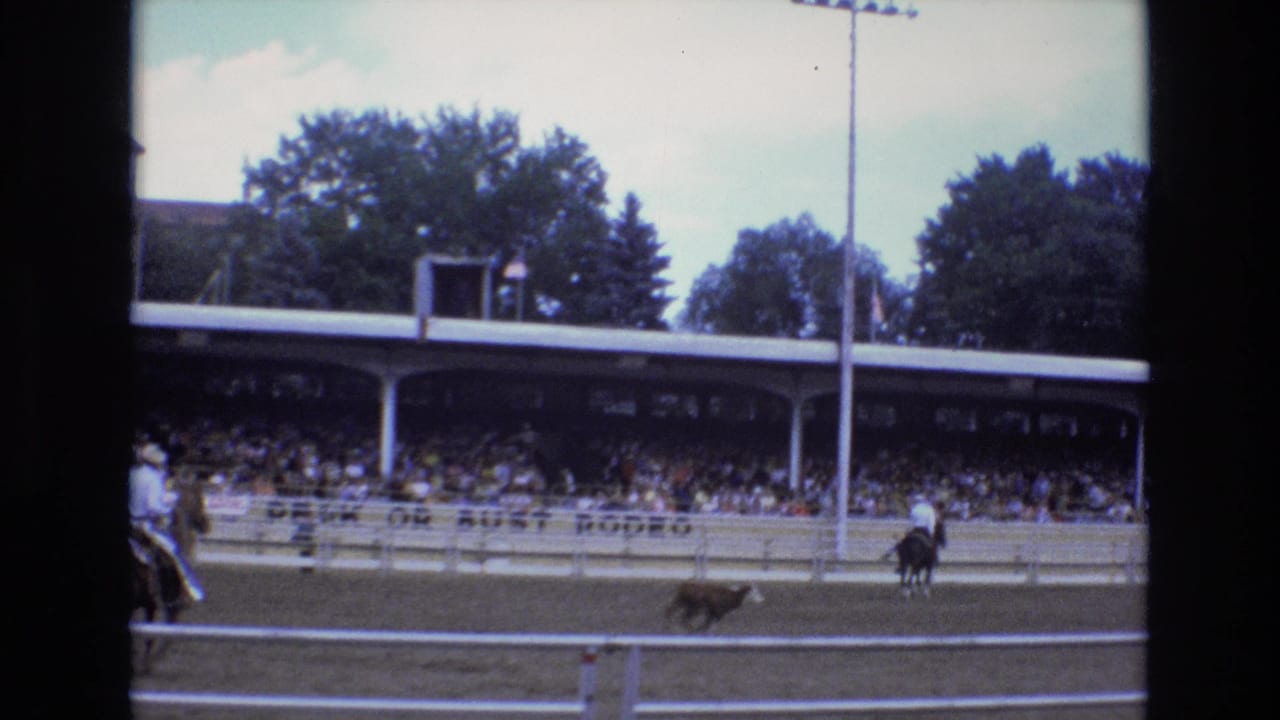 1973 Colorado Springs rodeo footage shows a mounted cowboy pursuing a calf in front of covered spectator bleachers and arena fencing.