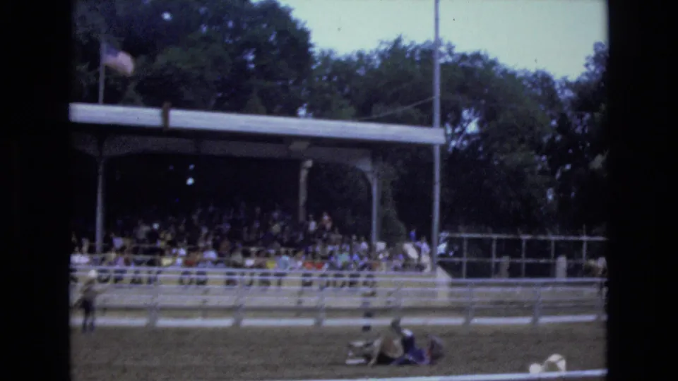 The home-movie frame shows the rodeo arena from the stands, with spectators watching and a figure low on the dirt near the rail during the roping sequence.