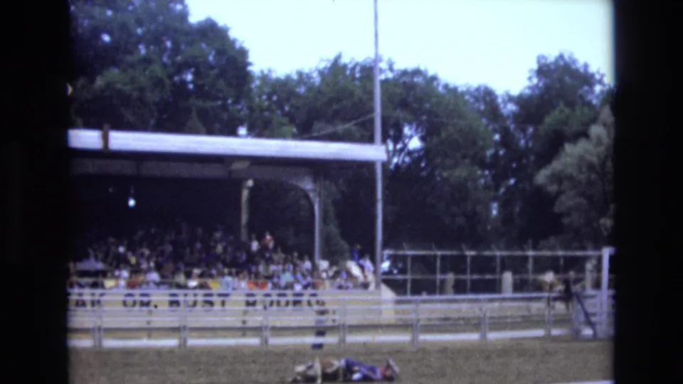 A later frame holds on the Colorado Springs rodeo arena, showing shaded spectators, fencing, trees, and a participant low on the dirt after the roping action.