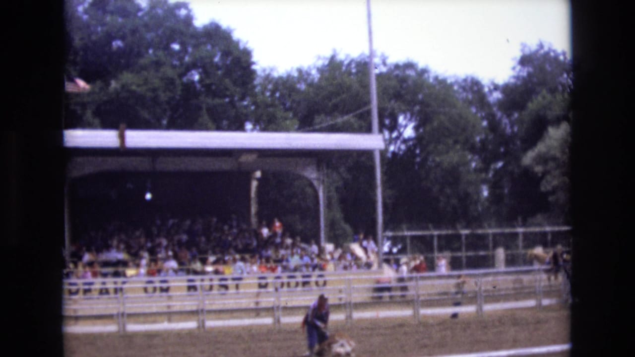 1973 Colorado Springs 8mm frame showing a racetrack grandstand filled with spectators, track railings, trees, and a partial American flag at the left edge.