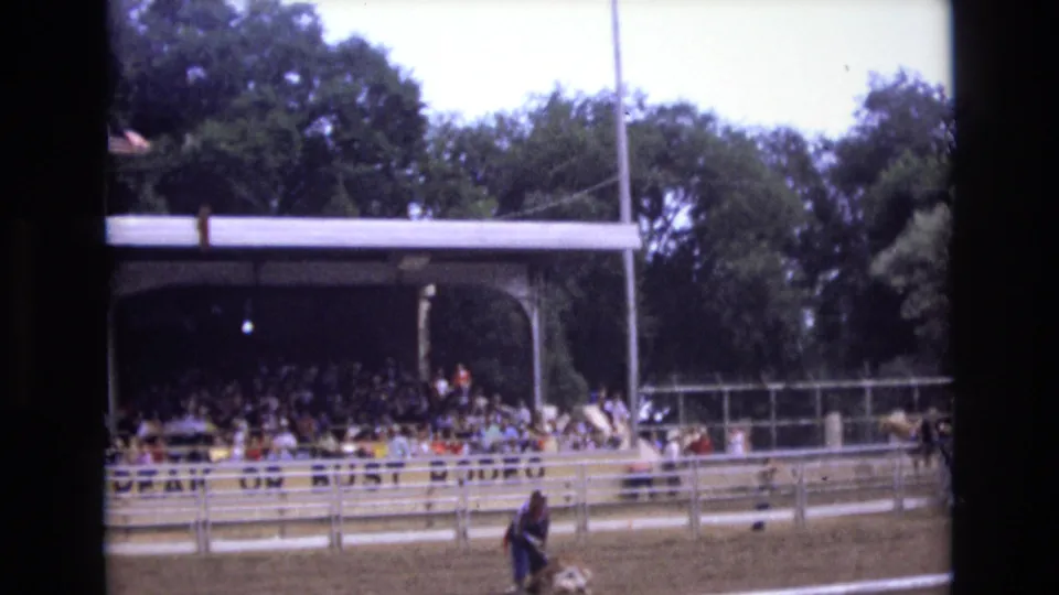 1973 Colorado Springs 8mm frame showing a racetrack grandstand filled with spectators, track railings, trees, and a partial American flag at the left edge.