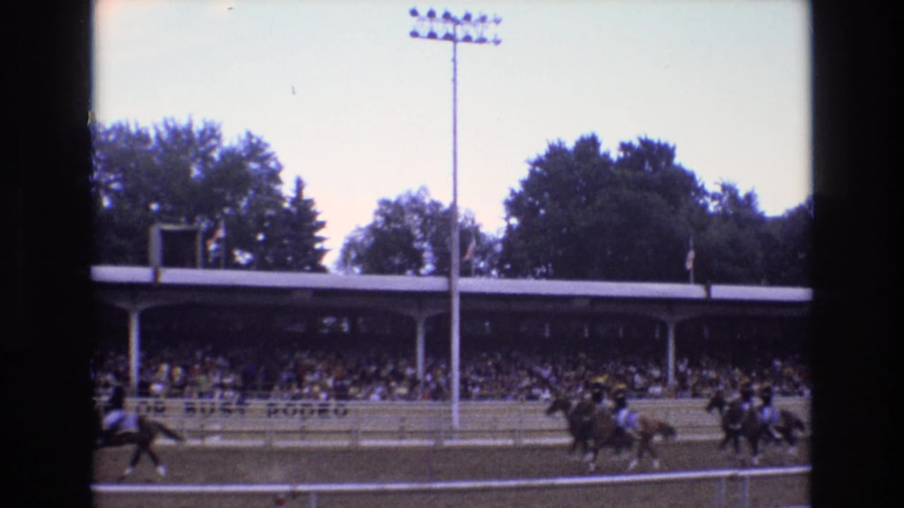 Racehorses and riders pass in front of a covered grandstand and tall stadium light.