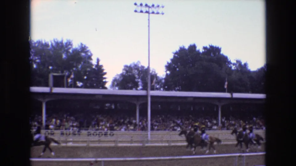 1973 Colorado Springs race-day frame with horses and riders moving along the dirt track before a covered grandstand and stadium light.