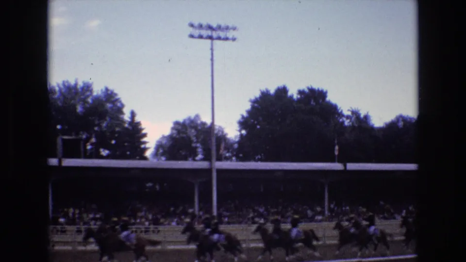 1973 Colorado Springs 8mm frame showing a group of horses and riders racing past the grandstand under a tall stadium light.