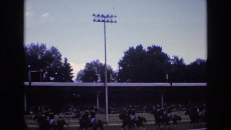 1973 Colorado Springs race-track frame with horses and riders moving past a shaded grandstand, trees, railings, and a tall stadium light.