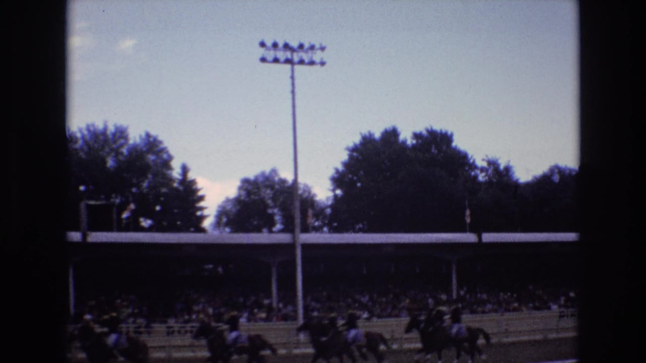 1973 Colorado Springs home-movie frame showing riders on horseback passing a dirt track grandstand with spectators, trees, and a tall stadium light pole in view.