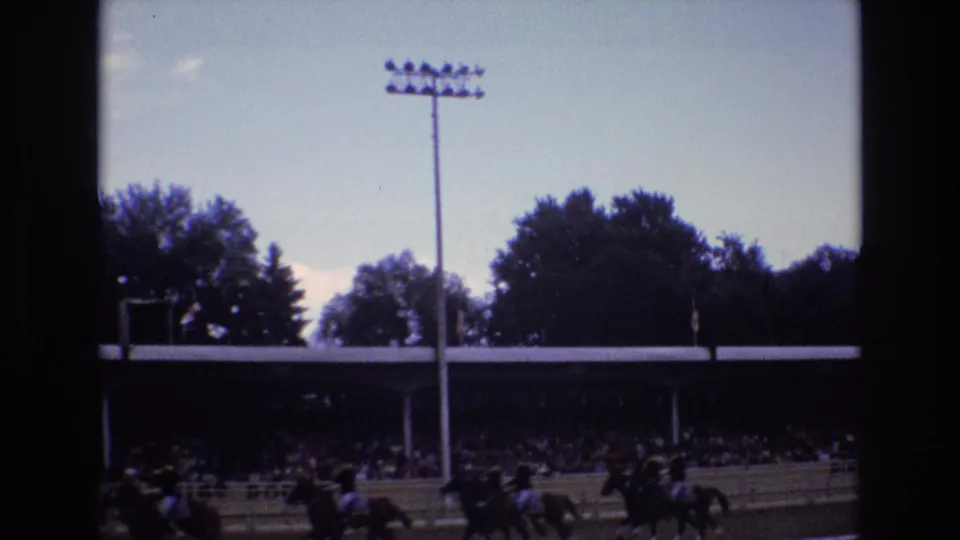 1973 Colorado Springs home-movie frame showing riders on horseback passing a dirt track grandstand with spectators, trees, and a tall stadium light pole in view.